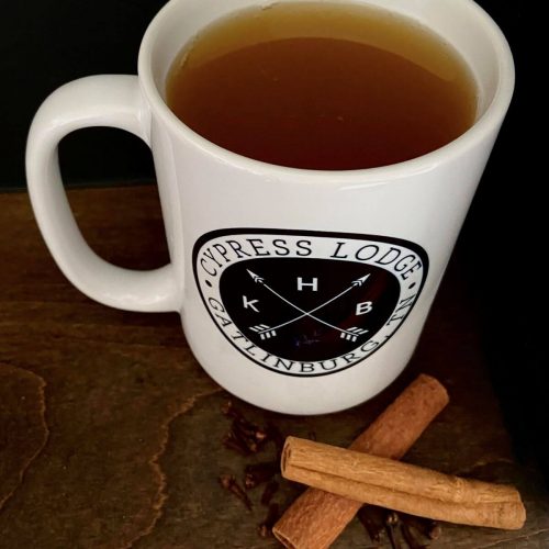 KHB Cypress Lodge Mug filled with warm peanut butter whiskey apple cider, a few cinnamon sticks and spices laying on the table beside the mug